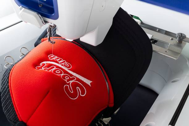 Embroidery machine and embroidered sport cap close up image Close up picture of white embroidery machine embroidering logo on red and black sport cap embroidery photos stock pictures, royalty-free photos & images