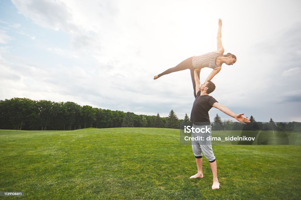 Equilibrio De Vida Pareja Deportiva Activa Haciendo Acro Yoga En La Naturaleza Un Hombre Fuerte Parado Sobre Una Hierba Y Equilibrando A Una Mujer En Su Brazo Foto de stock y más Equilibrio De Vida Pareja Deportiva Activa Haciendo Acro Yoga En La Naturaleza Un Hombre Fuerte Parado Sobre Una Hierba Y Equilibrando A Una Mujer En Su Brazo Foto de stock y más