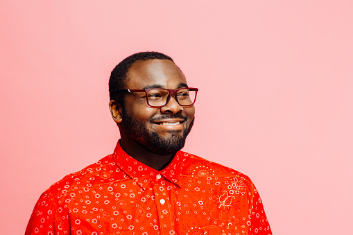 Man in bright red shirt and glasses  smiling and looking off camera against pink background