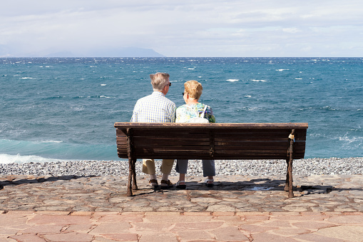Tenerife, Spain / November 2018. Elderly retired couple sitting on a bench, enjoying a sunny day by overlooking the Atlantic Ocean in the popular beach town Playa Las Americas on Tenerife, Spain.