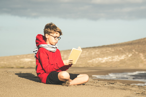 Cute boy in a red hoodie reading a book at the beach in a cloudy day, enjoying holiday. Young male sitting on the sand and relaxing with a blast. Kid enjoying a trip in a new exotic place near the sea
