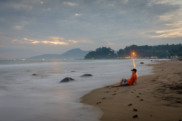 A man is sitting and enjoying the moment on Karang Hawu Beach, West Java, Indonesia with an orange sunset sky and blurry smooth waves