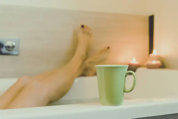 Photo of Crossed legs of woman in the water of the bathtub, girl relaxes immersed in a bath Relaxing candles atmosphere Rest and cool down after work moment for oneself Meditation cup coffee or herbal tea