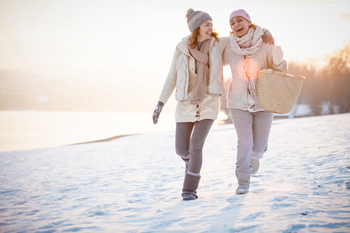 Two happy women are embracing while walking on snow-covered field on a sunny day.