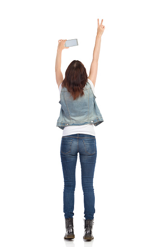 Rear view of young woman in jeans vest and black boots is standing, holding cell phone over her head, taking photo and showing peace hand sign. Full length studio shot isolated.