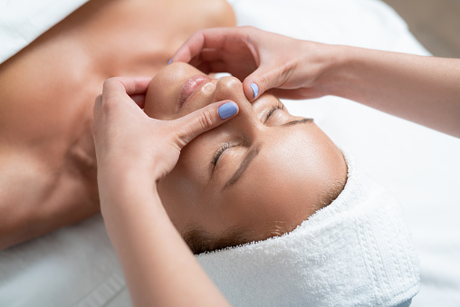 Feeling sleepy. Close up portrait of charming woman with closed eyes enjoying skincare procedure at spa salon. Masseuse hands with blue manicure touching client chin and nose