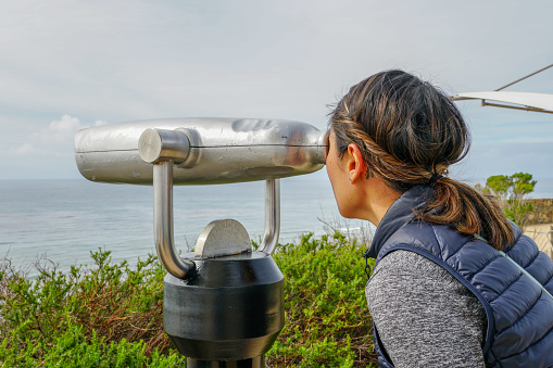 Asian woman looking in the binocular telescope on the tip of the Point Loma Peninsula in San Diego, California, USA. Young woman looking inside automated binoculars to observe SD city.