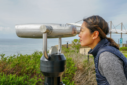Asian woman looking in the binocular telescope on the tip of the Point Loma Peninsula in San Diego, California, USA. Young woman looking inside automated binoculars to observe SD city.