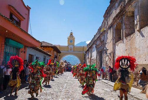 Antigua, Guatemala- December 2018: Locals and tourists in Antigua's street with famous arch and Agua volcano in the background watching a dance by dancers with traditional masks and costumes for the festivities to welcome the new year.