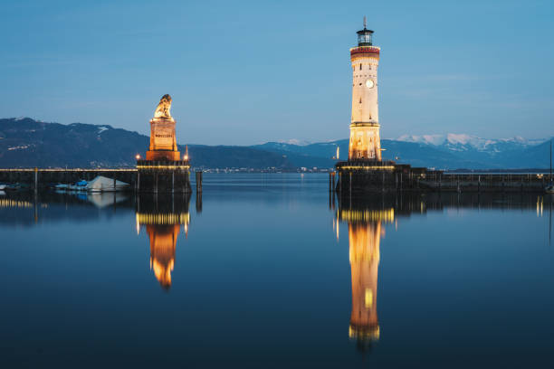 Lindau Lighthouse and Bavarian Lion at Night Bodensee Germany Illuminated Lindau Bodensee Harbor Entrance with the famous Lighthouse and Bavarian Lion Sculpture (from the year 1856) mirroring in the tranquil harbor pond at Night - Twilight. Snow covered European Alps in the background. Lindau, Bodensee, Bavaria, Germany. lindau lighthouse stock pictures, royalty-free photos & images
