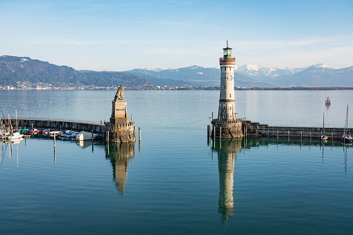 Aerial view towards Lindau Bodensee Harbor Entrance with the famous Lighthouse and Bavarian Lion Sculpture (from the year 1856) close to sunset, sun lit - snow covered european alps in the background. Lindau, Bodensee, Bavaria, Germany.