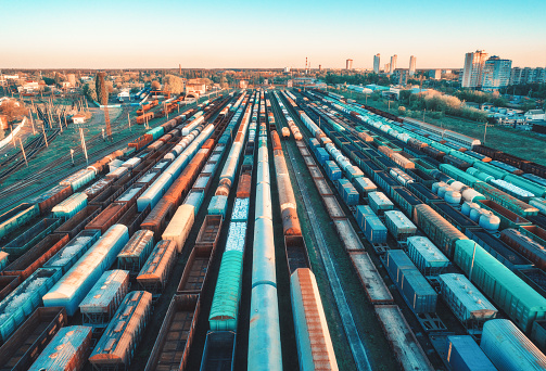 Aerial view of colorful freight trains on railway station at sunset. Wagons with goods on railroad. Heavy industry. Industrial scene with cargo trains, city buildings. Top view from drone. Vintage