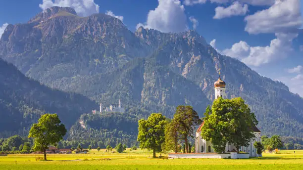 Bavarian landscape - view of the church of St. Coloman on the background of the Alpine mountains and Neuschwanstein Castle in summer day, Germany