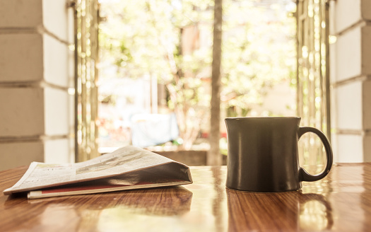 Cappuccino aroma cup of coffee for breakfast in cafe on wooden table background with newspaper. Morning sunlight coming from window on sun day or holiday. Leisure business idea. Copy space room text