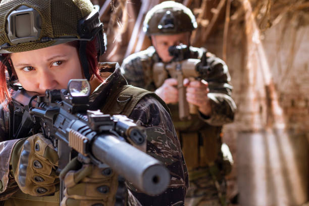 Female airsoft shooter aiming, male shooter standing behind. An old building with strong sunlight
