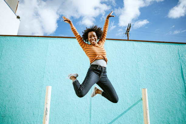 Celebrating Woman Jumps Into The Air A beautiful African American woman smiles, jumping high in the air with her arms outstretched and a smile on her face.  Horizontal image with copy space.  Shot in Los Angeles, California. jumping stock pictures, royalty-free photos & images