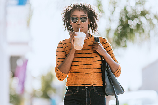 A young African American woman walks the streets of Los Angeles, a cold beverage in her hand.  Horizontal image with copy space.