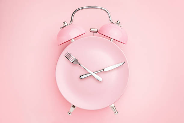 Alarm clock and plate with cutlery . Concept of intermittent fasting, lunchtime, diet and weight loss Alarm clock and plate with cutlery . Concept of intermittent fasting, lunchtime, diet and weight loss fasting activity stock pictures, royalty-free photos & images