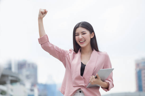 donna d'affari asiatica di successo braccia fino a festeggiare con tablet computer in mano in città all'aperto . ragazza eccitata vincitore. allegri, felice, donna che controlla online buone notizie - cheering arms raised women university foto e immagini stock