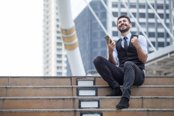 Cheerful Business Man Celebrating Success with mobile phone sitting on the stairs in urban city outdoors, looking up , good news,yes, raising hands,excited,Fulfill,winner , copy space stock photo