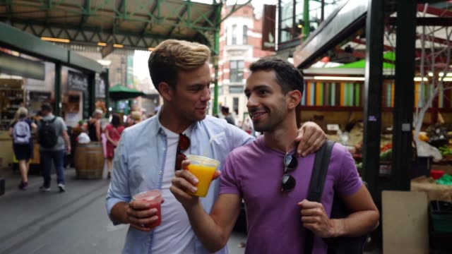 Happy gay couple walking through a street market talking and having fun and drinking juice