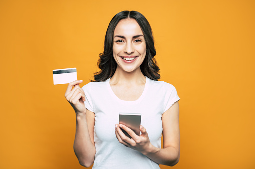 Check card. Bodacious girl with hazel eyes, slightly curled hair and such a brilliant smile in front of flamboyant orange background is holding her phone and a bank card.