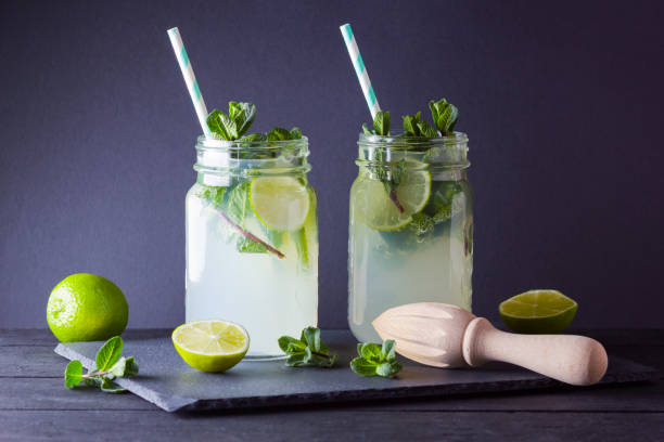 istock Mojito and mint on black background. Refreshing cocktail with lime and fresh mint on slate board. Two jars with handles with a summer drink with citrus and ice. Manual wooden juicer and mojito 1139430796