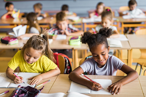 Happy Asian and African American elementary students writing in their notebooks while having a class at school. Their classmates are in the background.