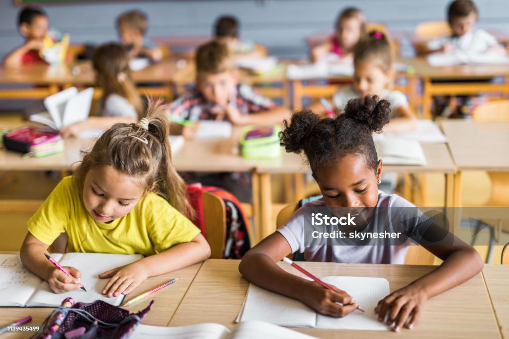 Happy schoolgirls writing a dictation on a class at school. Happy Asian and African American elementary students writing in their notebooks while having a class at school. Their classmates are in the background. Child Stock Photo
