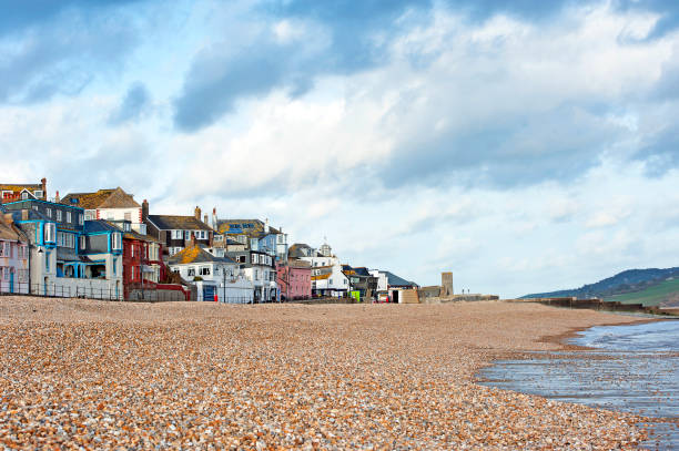 Colourful traditional houses and buildings of varying styles and periods almost reach the sand along the waterfront promenade walkway, or boardwalk, at low tide under an unsettled sky with moody clouds over a calm grey sea along the stony pebbles of the shingle beach of the Jurassic Coast coastline,  Lyme Regis, Dorset, England, UK