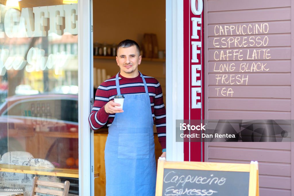 Portrait of small business coffee shop owner smiling and standing in front of shop serving a takeaway coffee, Successful young man working in trendy cafe store holding delicious cup of coffee 30-39 Years Stock Photo