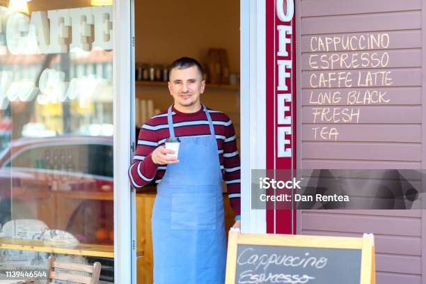 Portrait Of Small Business Coffee Shop Owner Smiling And Standing In Front Of Shop Serving A Takeaway Coffee Successful Young Man Working In Trendy Cafe Store Holding Delicious Cup Of Coffee Stock Photo - Download Image Now