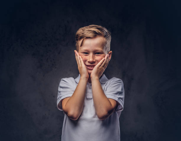 istock Surprised cute boy dressed in a white t-shirt stands with hands on face in a studio. Isolated on dark textured background. 1139453202