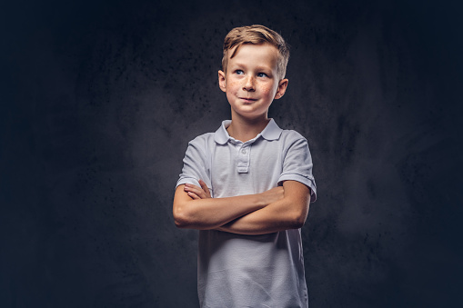 Cute little boy dressed in a white t-shirt standing with crossed arms in a studio. Isolated on a dark textured background.