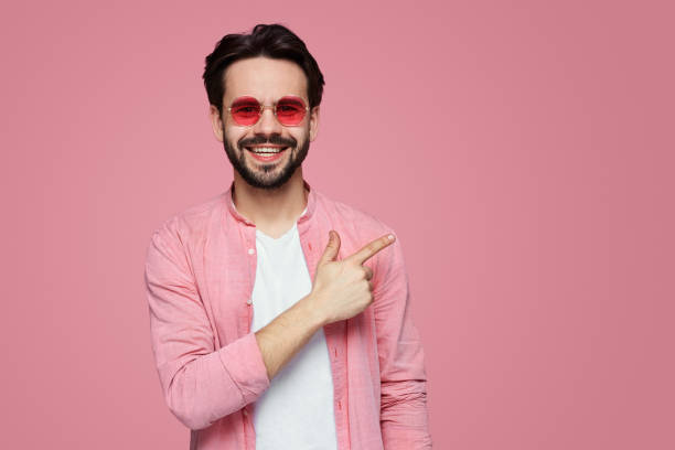 istock Hndsome young bearded boy in stylish shirt at copy space on pink wall points aside with cheerful expressionl. 1139460559