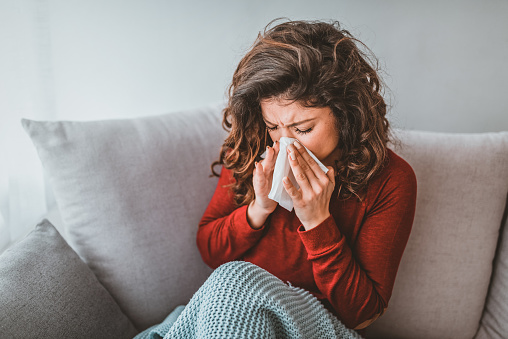 Portrait of woman with allergy blowing her nose. Illness And Sickness. Runny nose. Ill young blond woman having fever and blowing her nose while having a blanket. Portrait of a young woman sneezing in to tissue
