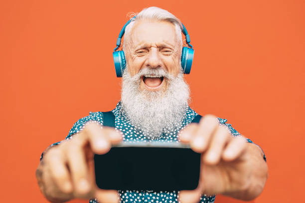 senior bearded mann macht selfie mit handy beim hören seiner lieblings-playlist mit kopfhörern-mode gereift männlich spaß mit smartphone-apps im freien-elderly, technologie-konzept - headphones music mobile phone tattoo stock-fotos und bilder