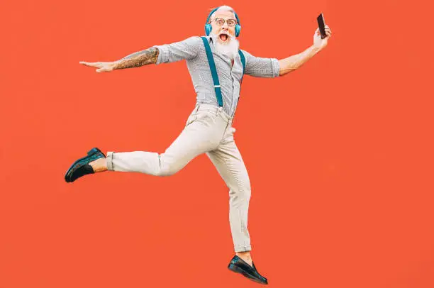 Photo of Senior crazy man jumping while listening music outdoor - Hipster male having fun dancing and celebrating life outside - Happiness, technology and elderly lifestyle people concept