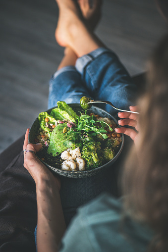 Healthy dinner or lunch. Curly woman in t-shirt and jeans sitting at home and eating vegan championship game or Buddha bowl with hummus, vegetable, fresh salad, beans, couscous and avocado, top view