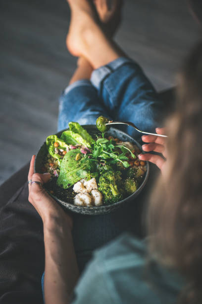Woman sitting at home and eating vegan championship game Healthy dinner or lunch. Curly woman in t-shirt and jeans sitting at home and eating vegan championship game or Buddha bowl with hummus, vegetable, fresh salad, beans, couscous and avocado, top view foxys_forest_manufacture stock pictures, royalty-free photos & images