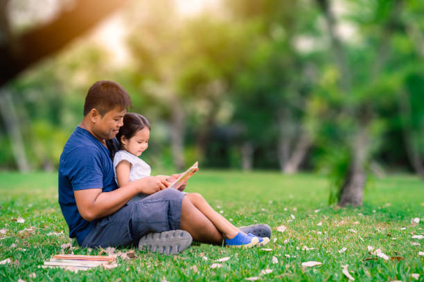 ojciec siedzi na trawie z córeczką i czyta książkę w parku - asian ethnicity male park men zdjęcia i obrazy z banku zdjęć
