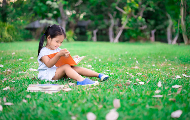маленькая милая девочка в синем платье, читающая книгу сидя в парке - little girls childhood outdoors horizontal стоковые фото и изображения