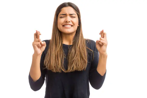 Woman having pleading expression on face while crossing fingers on white background