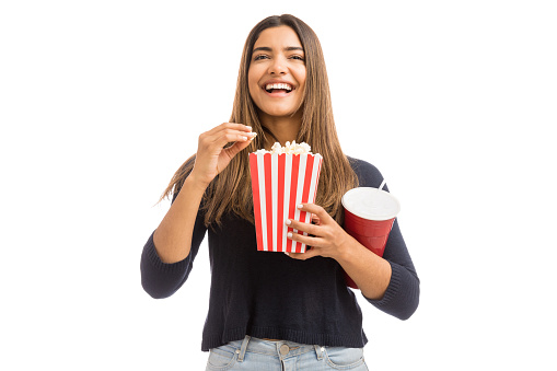 Cheerful Hispanic woman watching comedy movie while having snacks on plain background