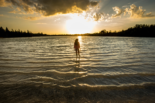 A beautiful woman wades into the water of a remote lake.  She has the wind in her hair and looking into the setting sun.