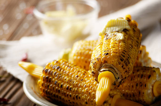 Wooden table with deep grilled sweet corn cobs under melting butter with plastic holder on clay dish
