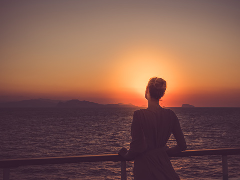Cute woman in fashionable dress on the top deck of a cruise ship, looking into the distance on a sunset background. Concept of sea cruises and rest