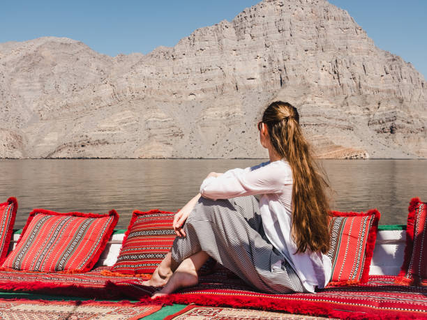 Fashionable woman sitting in a boat. Oman Fjords Fashionable woman sitting on a red blanket in a boat and looking into the distance on the background of coastal rocks. Oman Fjords. Concept of leisure and travel arabian sunglasses stock pictures, royalty-free photos & images
