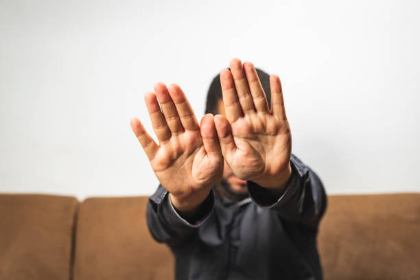 homem novo sério que senta-se no sofá e que mostra o gesto do batente-gesto da mão esconder o sinal da cara - patronize - fotografias e filmes do acervo