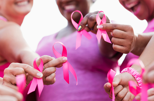 Cropped view of a multi-ethnic group of women of mixed ages standing together outdoors, wearing pink, at a breast cancer awareness rally, raising money to find a cure. They are each holding a breast cancer awareness ribbon. The focus is on their hands.